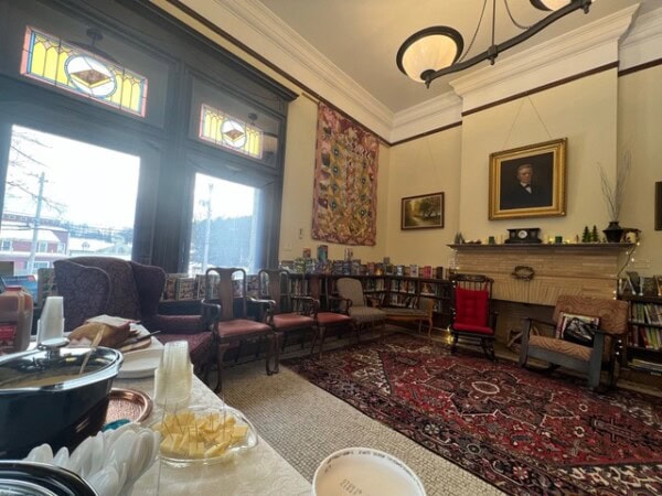a cozy library room with stained glass windows, assorted chairs, bookshelves, a fireplace, and a portrait above the mantle. a table with snacks is visible in the foreground.