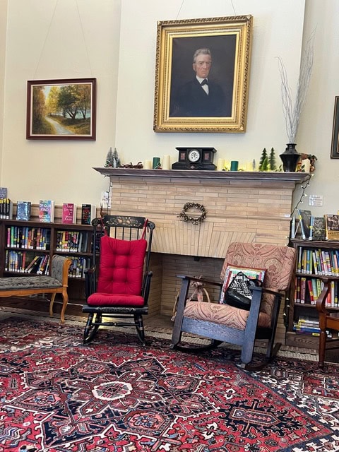 a library reading area with two rocking chairs, a fireplace, bookshelves, a patterned rug, and a portrait hanging above the mantel.