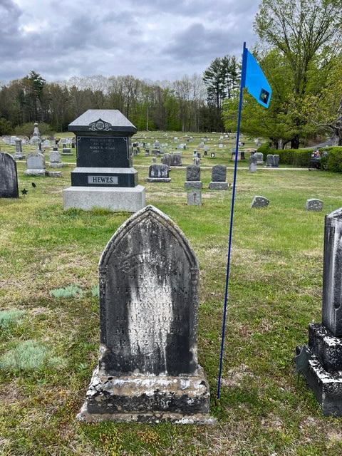 an old gravestone with worn text stands beside a blue flag in a cemetery, with more gravestones and trees visible in the background under a cloudy sky.