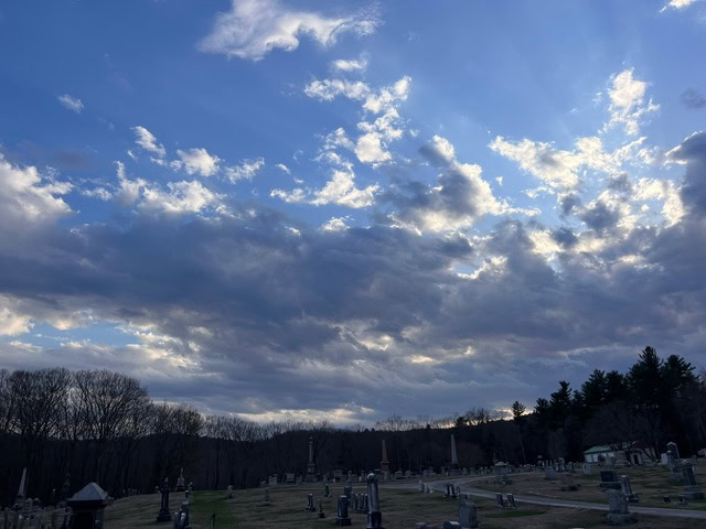 a cemetery with tombstones is shown under a dramatic sky with scattered clouds and rays of sunlight breaking through.