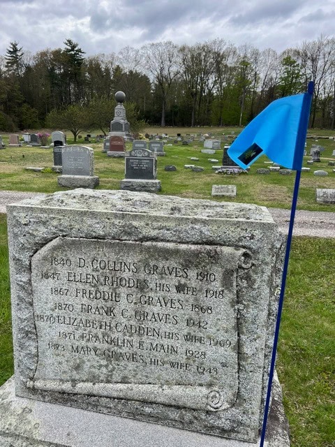 a gravestone with a blue flag in front, displaying names and dates of multiple individuals, set in a cemetery with other gravestones and trees in the background.