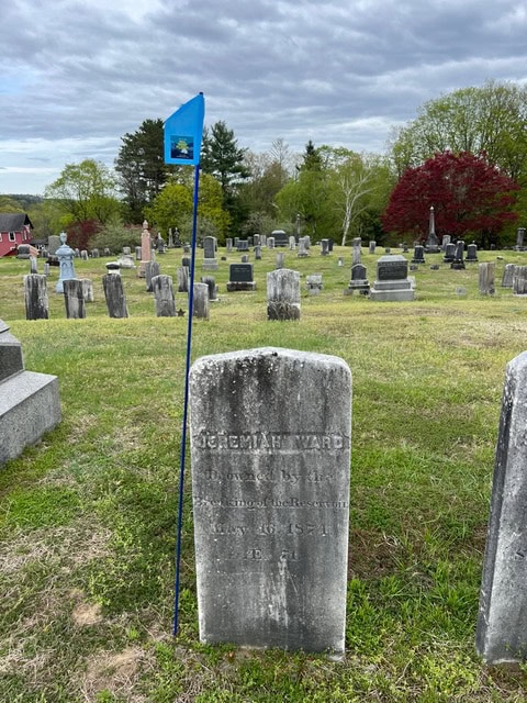 a blue flag marks the weathered gravestone of jeremiah ward in a grassy cemetery with other headstones and trees in the background.