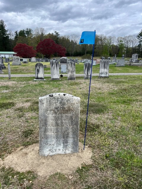 a gravestone stands in a cemetery with a blue flag on a pole placed beside it; other gravestones and trees are visible in the background under a cloudy sky.