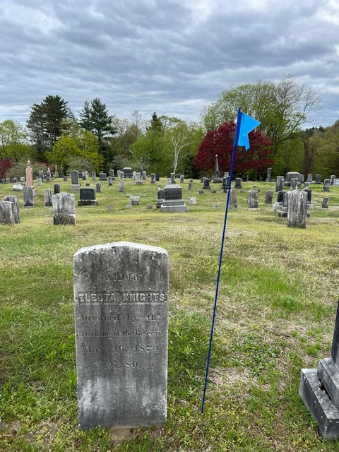 a weathered gravestone marked “electa knights” stands in a grassy cemetery, next to a blue flag on a pole, with more gravestones and trees in the background under a cloudy sky.