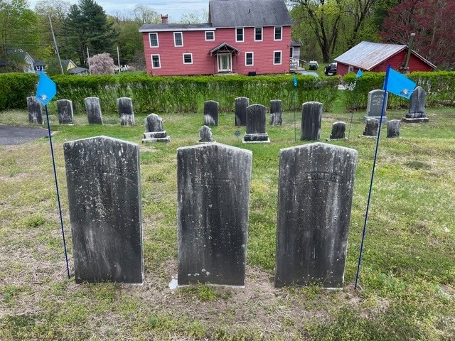 three old, weathered gravestones are lined up in front of several others, bordered by small blue flags, with a red house and trees in the background.