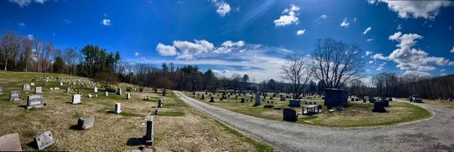 panoramic view of a cemetery with numerous headstones, a gravel road, scattered trees, and a bright blue sky with clouds.