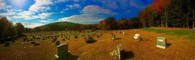 panoramic view of a cemetery with numerous headstones, surrounded by trees with autumn foliage under a partly cloudy sky.