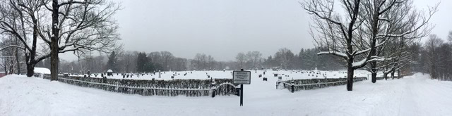 a snow covered cemetery with gravestones and leafless trees, surrounded by a fence and a central warning sign, under an overcast sky.