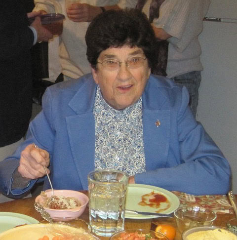 elderly woman in a blue jacket sits at a dining table with food, holding a spoon and smiling at the camera.