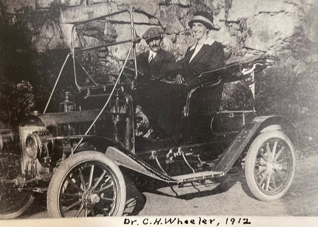 two people sit in an early 20th century open top automobile. the handwritten caption below reads: "dr. c.h. wheeler, 1912.