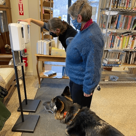 two women, both wearing masks, use a hand sanitizer station in a library. a german shepherd dog stands beside them. bookshelves and books are visible in the background.