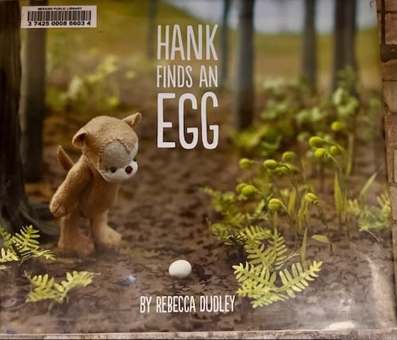 a stuffed animal character named hank looks at an egg on the ground in a forest setting. the book cover reads "hank finds an egg" by rebecca dudley.