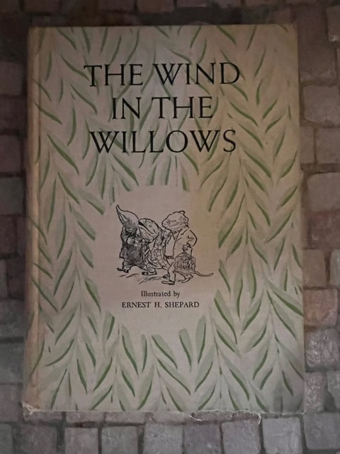 a worn cover of "the wind in the willows," illustrated by ernest h. shepard, featuring a small drawing of three animals and a leafy border pattern.