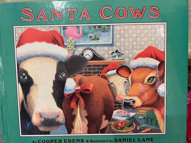 three cows wearing santa hats, one with a red bow, stand in a festive living room with a clock, christmas decorations, and gifts behind them. the book title is "santa cows.
