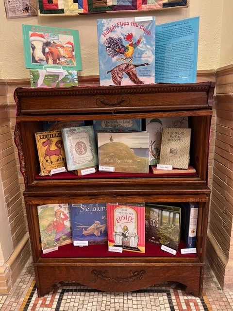 a wooden display shelf in a library holds a variety of children’s books with illustrated covers arranged on three tiers.