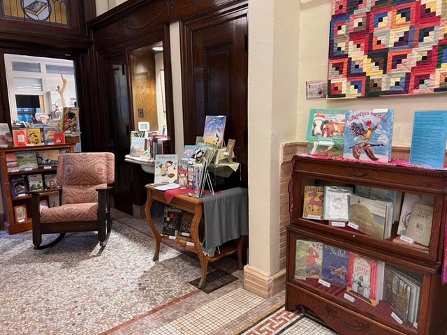 a reading area in a library with a cushioned chair, wooden shelves displaying books, a side table with more books, and a colorful quilt hanging on the wall.