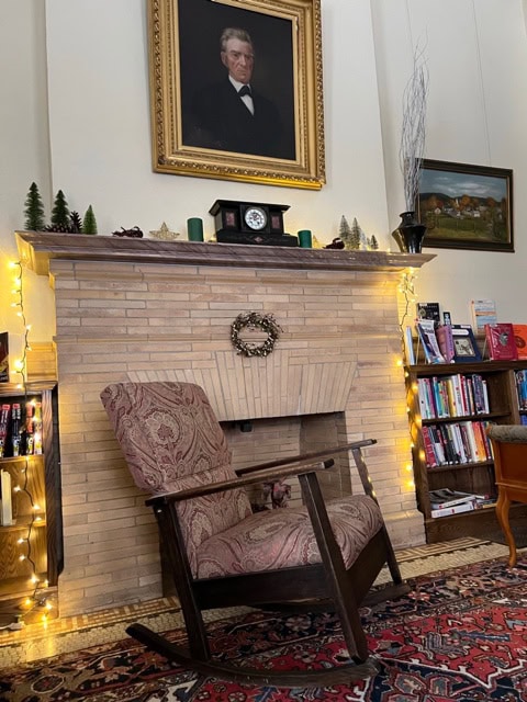 a patterned rocking chair sits in front of a fireplace with a wreath, bookshelves, string lights, and a portrait of a man above the mantel.