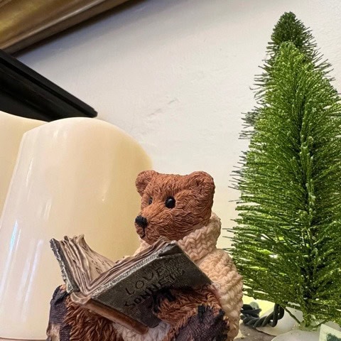 a small bear figurine wearing a sweater sits next to a miniature green christmas tree, holding an open book.