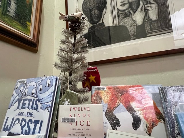 a small white christmas tree is displayed on a bookshelf with children's books, under a black and white drawing hanging on the wall.