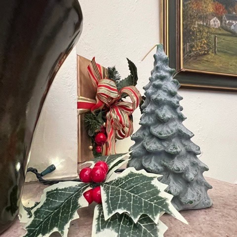 a ceramic christmas tree, artificial holly leaves with red berries, and a decorative ribbon are arranged on a shelf next to a painting and a large dark vase.