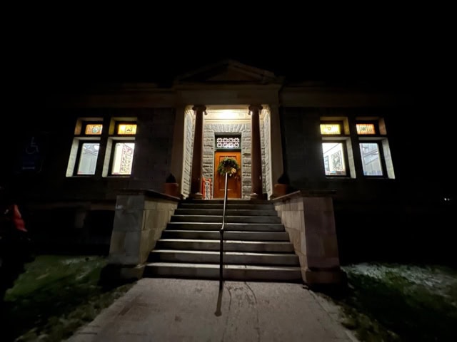 a stone building with lit stained glass windows and a central staircase leading to a wooden door, photographed at night.