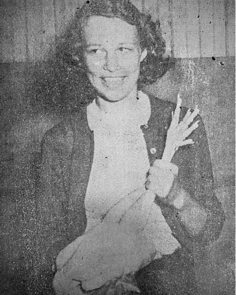 a woman sits indoors, smiling and holding a white chicken in her lap.