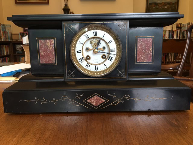antique black mantel clock with gold and red decorative panels, roman numeral face, and ornate detailing, placed on a wooden table in a library setting.