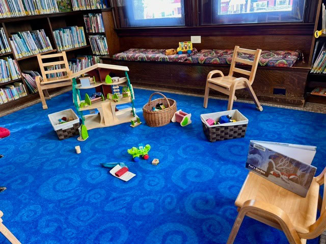 children’s play area in a library with wooden chairs, toys, books, baskets, and a blue carpet surrounded by bookshelves.