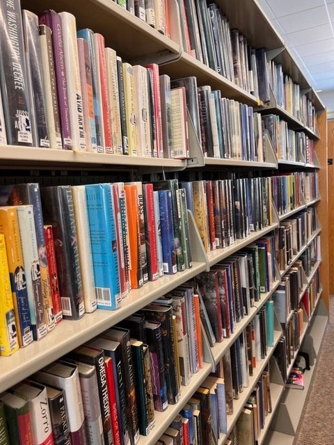 rows of books arranged on library shelves, with book spines facing outward, in a well lit room with a carpeted floor.