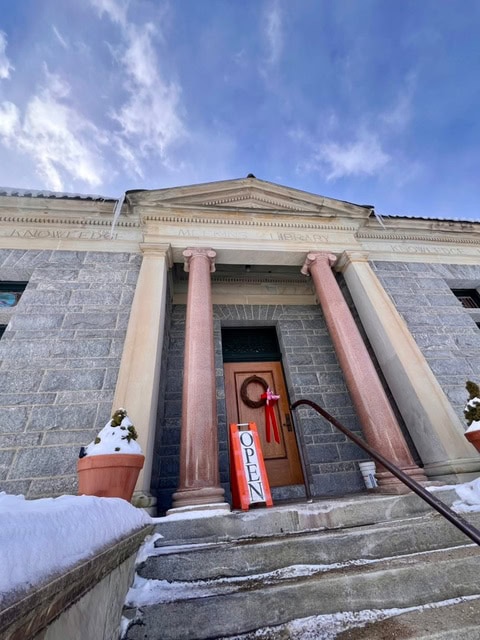 stone building with two large columns, snowy steps, a "open" sign by the door, and a wreath hanging on the entrance under a partly cloudy sky.