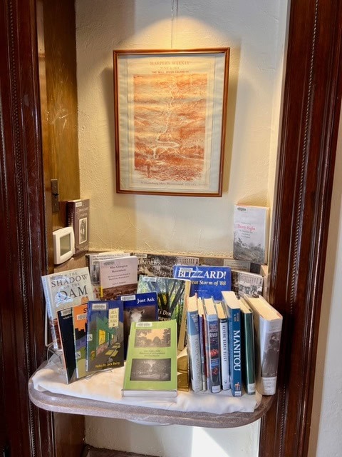 a small display nook with various books and pamphlets arranged on a shelf beneath a framed picture hanging on the wall.