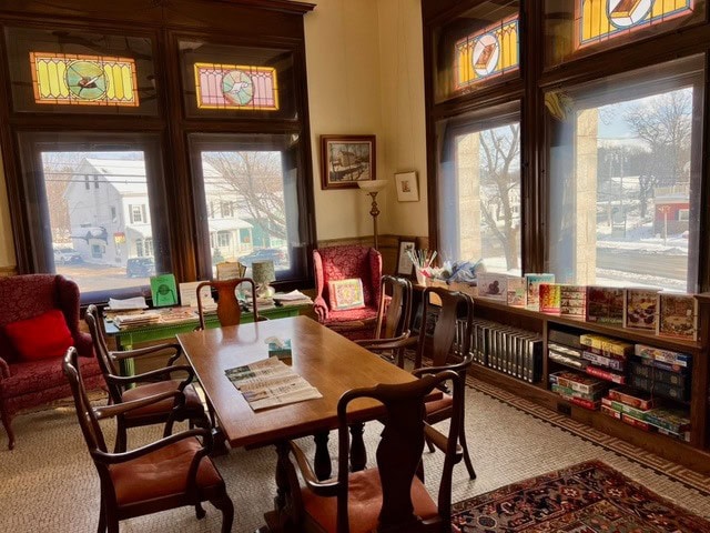 a cozy reading room with wooden table, chairs, armchairs, books, board games, large windows, and stained glass panels, with sunlight streaming in and a snowy scene visible outside.