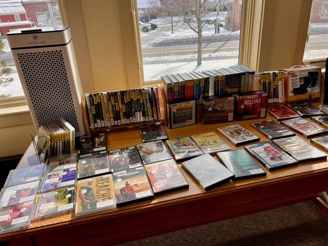 a wooden table covered with dvds, books, and audiobooks is positioned in front of windows overlooking a snowy street. an air purifier stands to the left of the table.