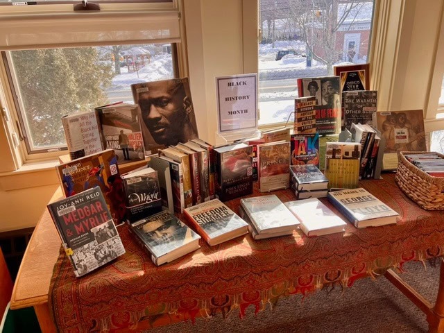 a table covered with books about black history and culture is displayed in front of windows, with a sign reading "black history month" in the center.