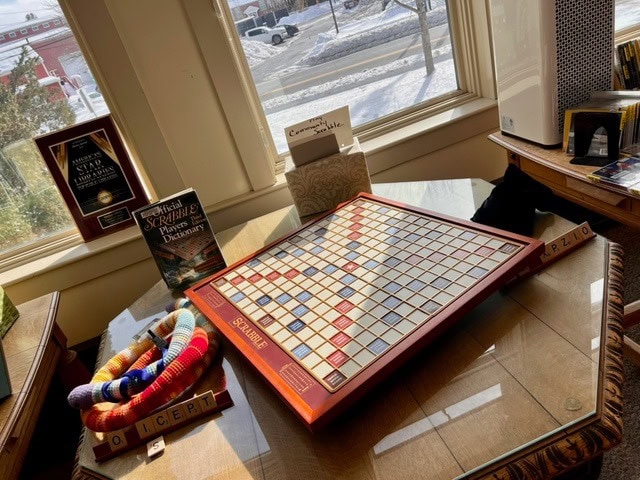 a scrabble board sits on a wooden table near a window, surrounded by a colorful scarf, letter tiles, plaques, and a scrabble dictionary, with snow visible outside.