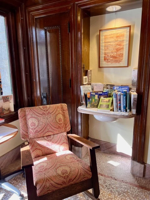 a cushioned armchair sits by a wooden door and window next to a small shelf holding books and pamphlets below a framed picture on the wall.