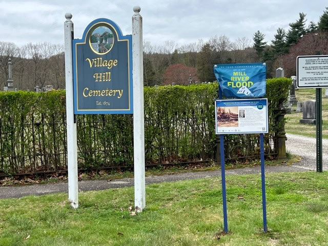 a sign reading “village hill cemetery est. 1741” stands next to informational displays about the mill river flood, with graves and trees visible in the background.