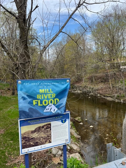 a blue sign commemorates the mill river flood beside a small river, with trees and a partly cloudy sky in the background.