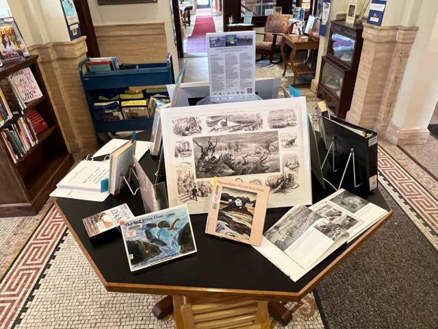 a library display table holds books, large black and white illustrations, and informational pamphlets, with bookshelves and seating visible in the background.