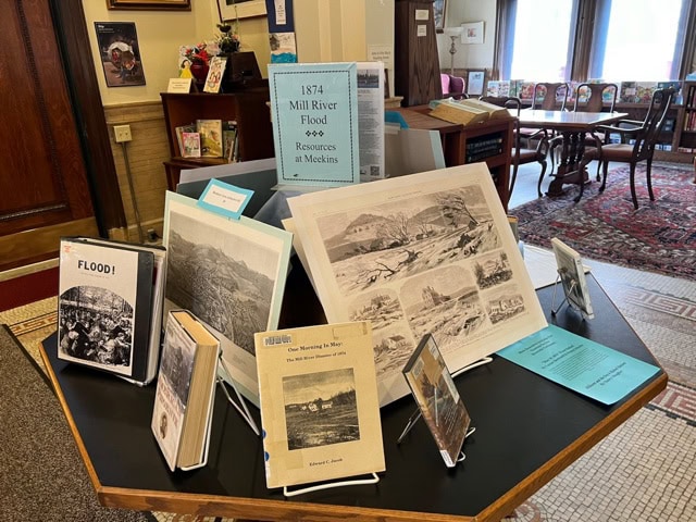 a display table with books, pamphlets, and illustrations about the 1874 mill river flood is arranged in a library setting near windows and bookshelves.