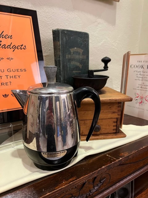 a stainless steel farberware percolator sits on a cloth covered surface next to a vintage wooden coffee grinder and old cookbooks.