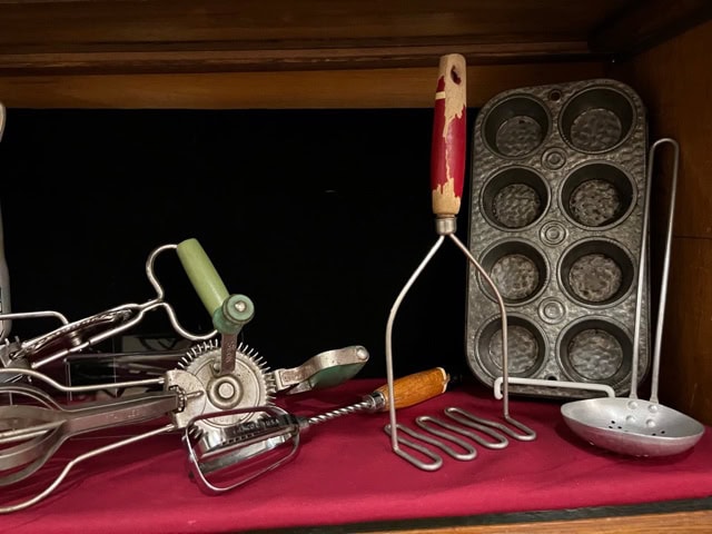 a collection of vintage kitchen tools, including a potato masher, muffin tin, hand mixer, and a slotted spoon, displayed on a red cloth.