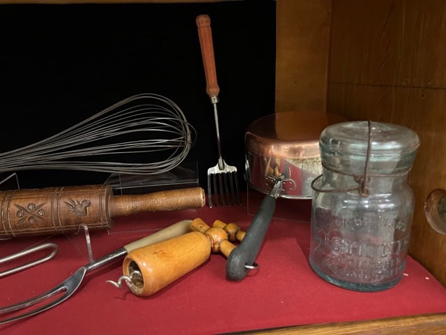 a display of vintage kitchen utensils including a whisk, rolling pin, jar, saucepan, potato masher, and various wooden handled tools on a red surface.