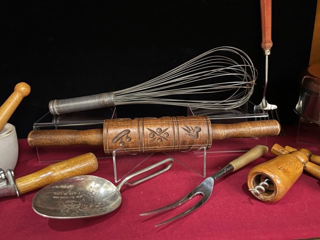 a collection of vintage kitchen utensils, including a patterned wooden rolling pin, metal whisk, meat fork, pastry blender, and scoops, displayed on a red cloth.