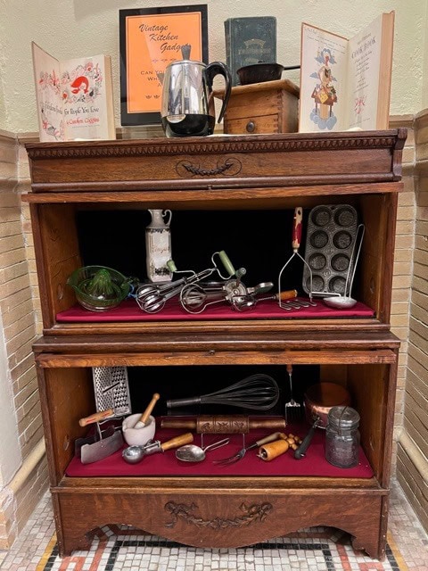 a wooden cabinet displays vintage kitchen tools, baking pans, utensils, an old cookbook, and a pitcher, all arranged neatly on red velvet fabric.