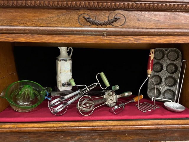 a collection of vintage kitchen tools including a juicer, hand mixers, a pitcher, a potato masher, a muffin tin, and a metal ladle displayed on a red cloth inside a wooden cabinet.
