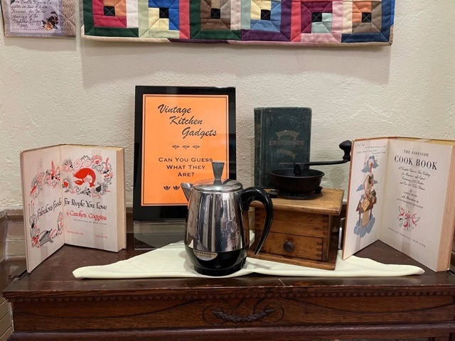 a display of vintage kitchen gadgets includes an old coffee grinder, a stainless steel percolator, two open cookbooks, and a sign reading "vintage kitchen gadgets" on a wooden table.