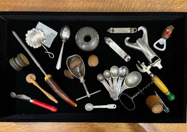 a collection of vintage kitchen tools and utensils, including measuring spoons, a corkscrew, a nutcracker, and various metal gadgets, arranged in a black tray.