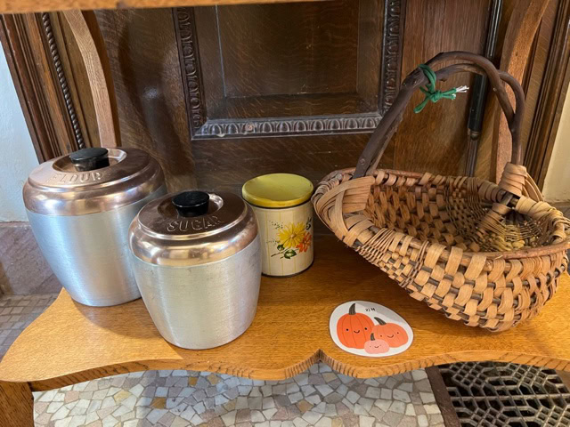 two metal canisters labeled "flour" and "sugar," a small ceramic container with a sunflower design, a wicker basket, and a sticker with pumpkins on a wooden shelf.