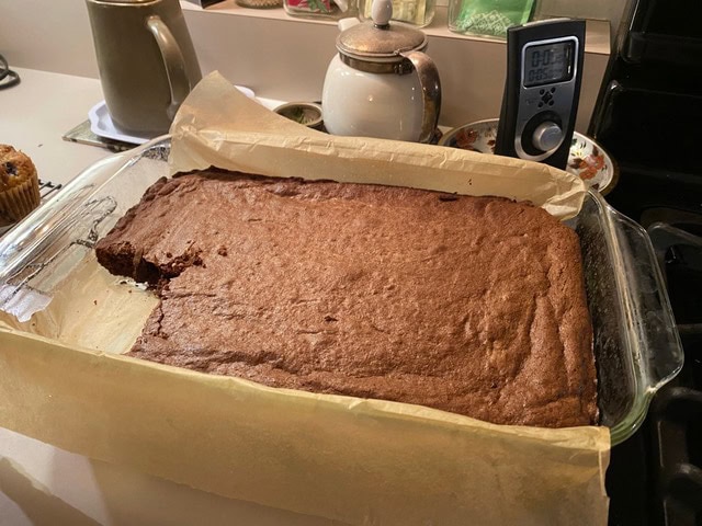 a rectangular pan of brownies with one piece missing, lined with parchment paper, sits on a kitchen counter near a teapot, timer, and other kitchen items.